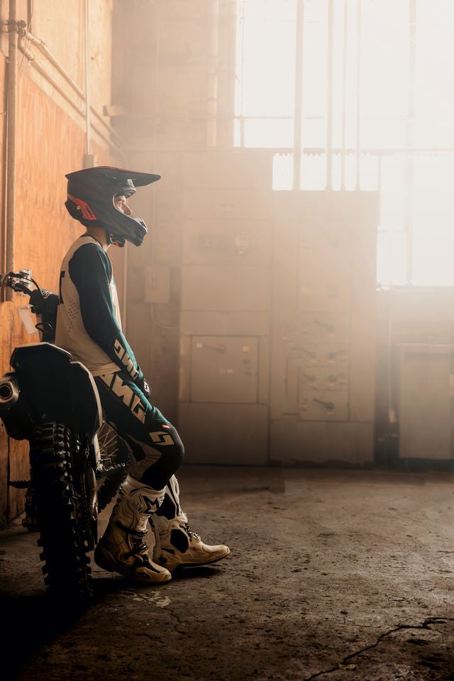 Person in motocross gear sitting on a dirt bike in a sunlit room with large windows.
